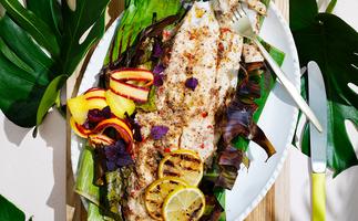 Aerial view of a barbecued coral trout wrapped partially in a banana leaf with grilled lemons and carrot escovitch. 