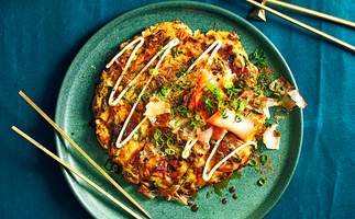 Overhead shot of a single okonomiyaki (Japanese pancake) on a green plate with chopsticks and blue cloth in frame